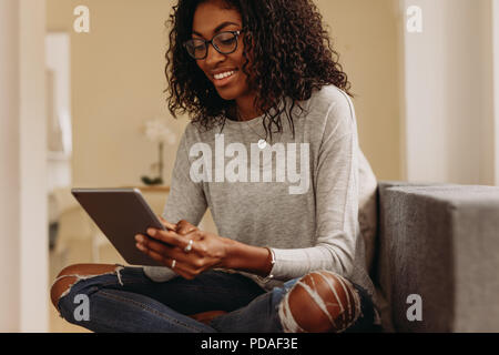 Woman sitting on sofa at home working on tablet pc. Smiling woman using a tablet pc while sitting at home with legs crossed. Stock Photo