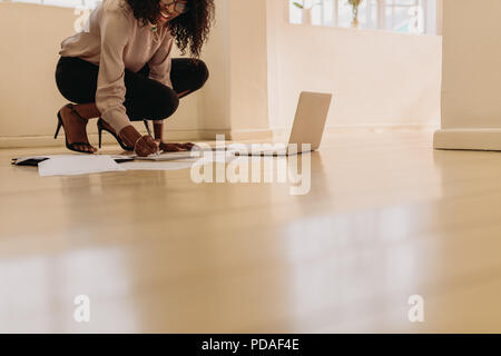 Woman entrepreneur in formal attire making notes while working on laptop computer at home. Businesswoman sitting on floor wearing sandals at home maki Stock Photo
