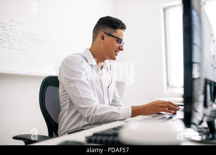 Smiling software developer sitting at his office desk working on laptop wearing earphones. Man wearing spectacles working on laptop computer in office Stock Photo