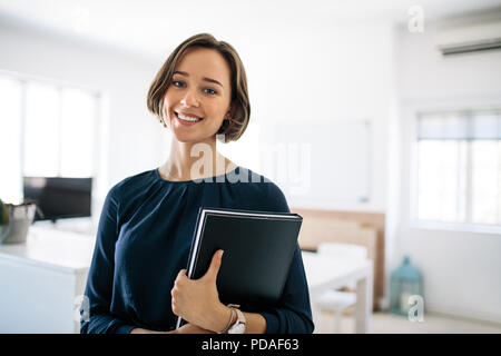 Smiling businesswoman standing in office holding a diary. Portrait of a happy woman entrepreneur at work. Stock Photo