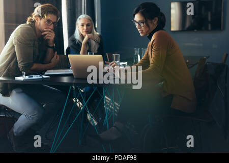 Three business people sitting around a table and looking at laptop. Business woman showing project analysis on laptop to colleagues. Stock Photo