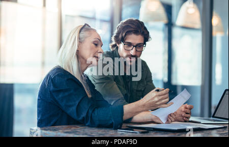 Mature woman and young man discussing work in office. Businesswoman holding a paper in hand while taking to her male colleague. Stock Photo