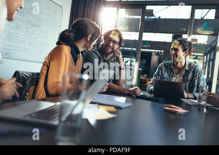 Business team sitting around a table in office lobby with sticky notes on their forehead having a brainstorming meeting. Corporate business people sit Stock Photo