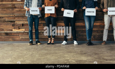 Low section of business people standing in row against a wall holding names of their respective departments on a placard. Candidates shortlisted of an Stock Photo