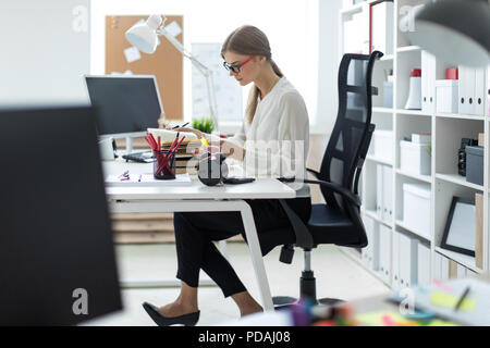 A young girl sits at a table in the office and holds a yellow marker in her hand. Before the girl lies an open book. Stock Photo