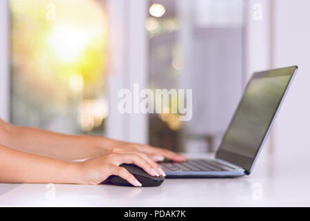 Beautiful female hand holding computer mouse and typing on laptop keyboard in office. Modern work and job concept. Sunset and window blurred Stock Photo