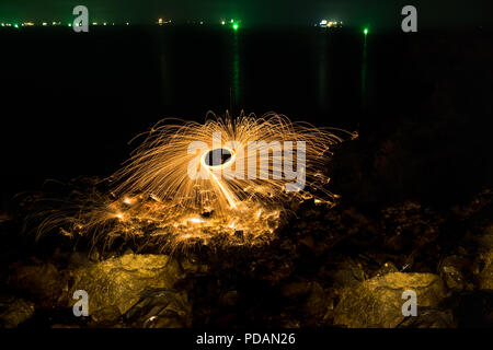 Man showers of glowing sparks from spinning steel wool on rocky beach at night. Stock Photo