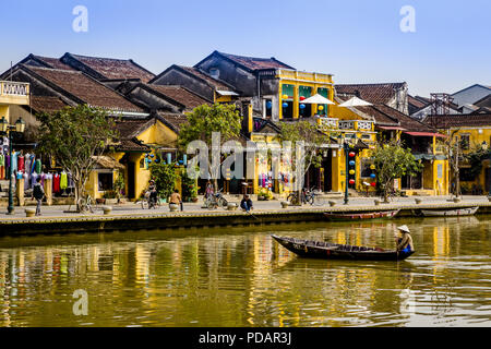 River front in Hoi An with a boatman approaching the water front with his small boat. The yellow building and colors permeate the scene. Stock Photo