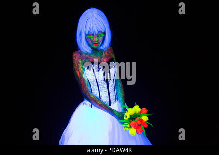 concept of wedding photosession in neon lighting in vogue style bride holds flower bouquet glows in the dark young girl with hairstyle quads and bouquet posing in white wedding dress under the UV rays on a dark background in the studio Stock Photo