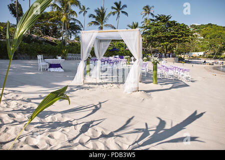 Beach wedding venue settings at seaside on the white sand, Arches and altar decoratiing with white soft fabric on the bamboo frame, white chiavari cha Stock Photo