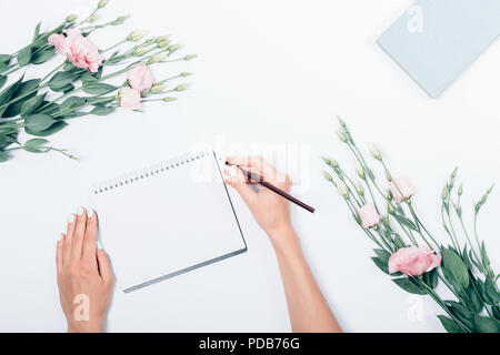 Woman's hand with pencil making notes on blank sheet near book and bouquet of flowers, top view. Flat lay composition stylized feminine workplace. Stock Photo