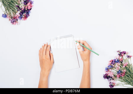 Flat lay floral mock-up of woman making notes on blank sheet. Female hands writing diary among pink and green flowers on white background, top view. Stock Photo
