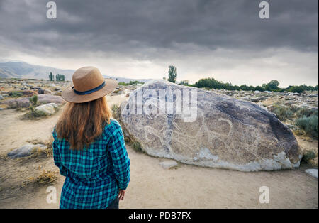 Woman in Hat in blue shirt in the open air museum near stone with ancient petroglyph of goats at Kyrgyzstan. Culture Heritage Museum. Stock Photo