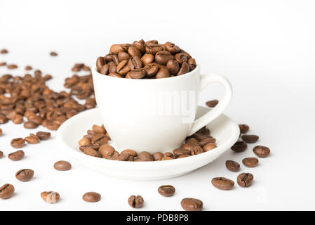 White china espresso cup on white background filled with roasted coffee beans which have spilled onto surface Stock Photo