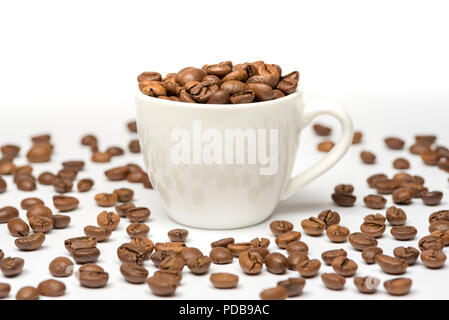 White china espresso cup on white background filled with roasted coffee beans which have spilled onto surface Stock Photo