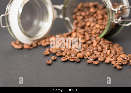 Glass food storage jar lying on side with coffee beans spilling out on black surface Stock Photo
