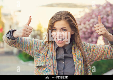 Bright young woman looking super excited while holding thumbs up in approval and smiling at camera outdoors Stock Photo