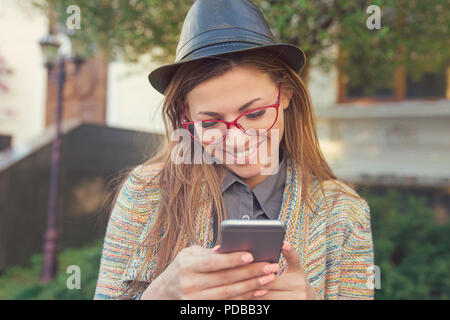Trendy modern hipster woman in hat and glasses chatting via smartphone and smiling while standing on street