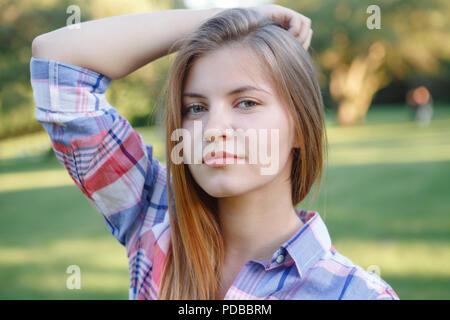 Closeup portrait of beautiful smiling young Caucasian woman with long red hair in plaid shirt posing in park at summer sunset, looking in camera Stock Photo