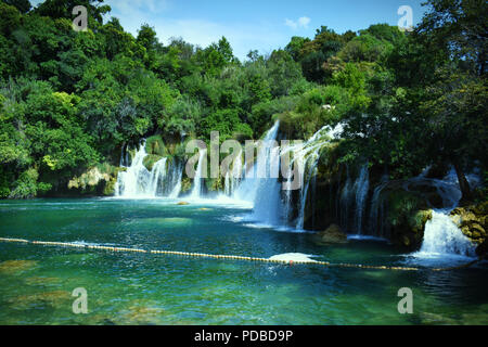 Beautiful Clear Turquoise Waterfalls Leading into Large Pool of Water that is Surrounded by a Green Forrest with Trees and Blue Sky with Fluffy Clouds Stock Photo