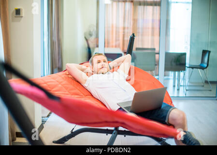 Young man with laptop holding hands behind head, relaxing and dreaming while lying on a hammock in office space. Taking time for a minute break. Unpro Stock Photo