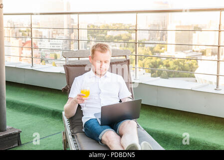 Portrait of Young businessman with drink working on laptop comfortably sitting on deckchair on office terrace with Urban Landscape on Background. Casu Stock Photo