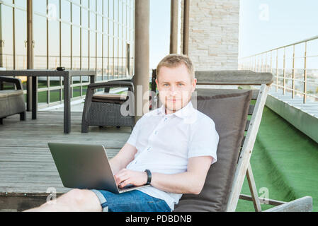 Portrait of Young smiling businessman working on laptop comfortably sitting on deckchair on office terrace with Urban Landscape on Background. Casual  Stock Photo