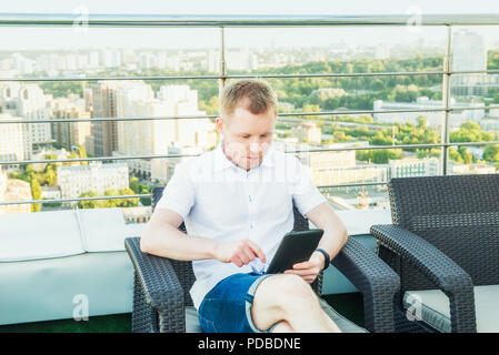 Young businessman working on laptop tablet comfortably sitting on chair on the office terrace balcony with Urban Landscape on Background. Casual Cloth Stock Photo