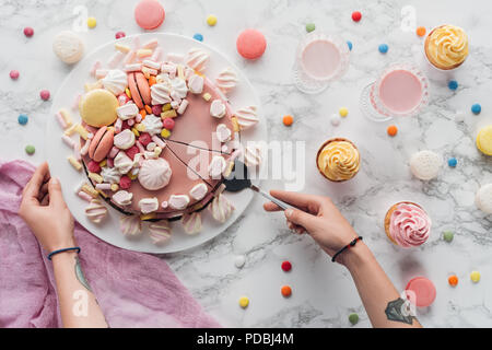 cropped view of woman taking piece of pink birthday cake with marshmallows and macarons Stock Photo