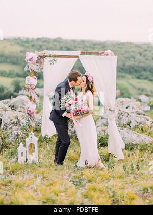 The full-length view of the kissing newlyweds near the lovely decorated arch during the wedding ceremony in the mountains. Stock Photo