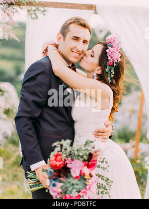 Close-up portrait of the bride kissing the smiling groom in the cheek during the wedding ceremony in the mountains. Stock Photo