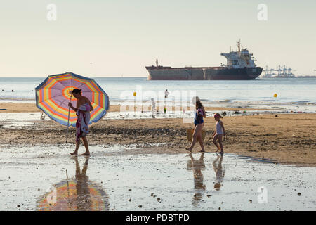 Holidaymakers walk along the beach, the Plage du Butin, at Honfleur as an oil tanker passes out to sea down the Seine Estuary by Le Havre Stock Photo