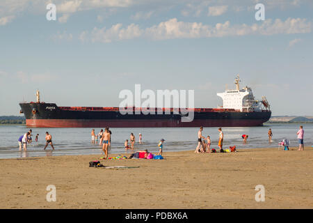 Holidaymakers walk along the beach, the Plage du Butin, at Honfleur as an oil tanker passes out to sea down the Seine Estuary by Le Havre Stock Photo