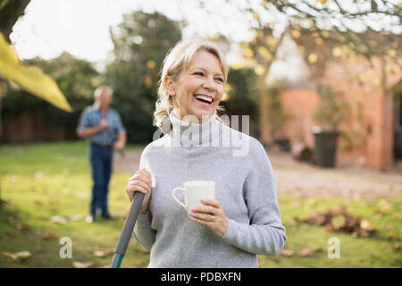 Happy mature woman drinking coffee and raking autumn leaves in backyard Stock Photo