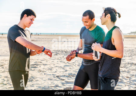 Male runners checking smart watch fitness trackers on sunny beach Stock Photo