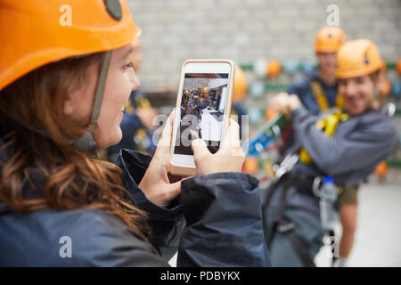 Woman with camera phone photographing friends in zip line equipment Stock Photo