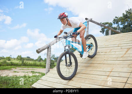 Focused mature man mountain biking down obstacle course ramp Stock Photo