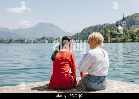 back view of stylish girls sitting on wooden pier near tranquil mountain lake, bled, slovenia Stock Photo