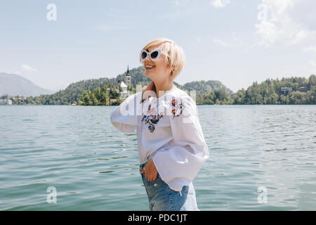 beautiful smiling young woman in sunglasses standing near majestic mountain lake, bled, slovenia Stock Photo