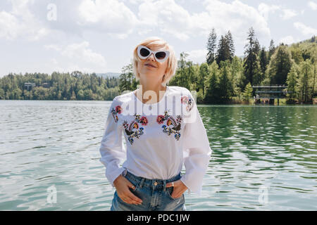 beautiful young woman in sunglasses standing near scenic tranquil mountain lake, bled, slovenia Stock Photo