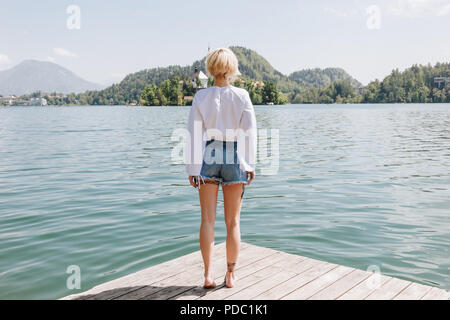 back view of young woman standing on wooden pier and looking at scenic mountain lake, bled, slovenia Stock Photo