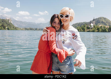 beautiful young women in sunglasses hugging and smiling at camera while standing near tranquil mountain lake, bled, slovenia Stock Photo