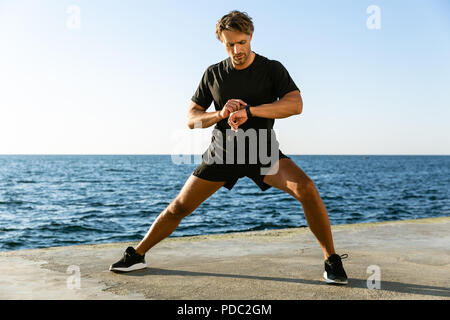 handsome adult sportsman looking at fitness tracker while stretching before training on seashore