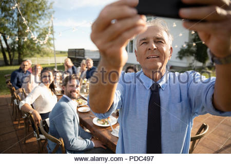 Senior man with camera phone taking selfie with friends at retirement party in sunny rural garden