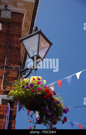 Bunting Against a Blue Sky at Sidmouth Folk Festival, by Wall Mounted Traditional Lamp and Hanging Basket Flowers. East Devon, UK. August, 2018. Stock Photo