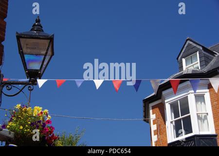 Bunting Against a Blue Sky at Sidmouth Folk Festival, by Wall Mounted Traditional Lamp and Hanging Basket Flowers. East Devon, UK. August, 2018. Stock Photo