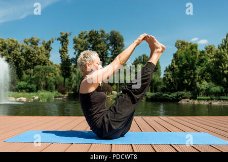 side view of woman practicing yoga and stretching legs on yoga mat near river in park Stock Photo