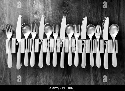 lots of knives forks and spoons staked in a line in five wooden cutlery holders on a wooden table top, black and white photograph Stock Photo