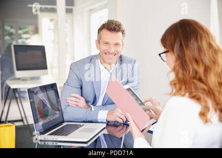 Businesswoman using her digital tablet while sitting at office desk with her colleague and having discussion. Teamwork at the office. Stock Photo