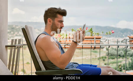 Waist Up Profile of Attractive Man with Dark Hair, Sitting with Tablet Computer Working or on His Start-up Business on a Terrace or Balcony in Summer  Stock Photo